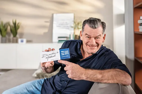 Mark Kühler auf einem Sofa mit einem Schild „Die starken Händler“ und Europa Möbel-Logo in der Hand – sympathische Markenbotschaft für EM-Kampagne, starke Möbelhändler, hochwertige Einrichtung und Vertrauen beim Möbelkauf.